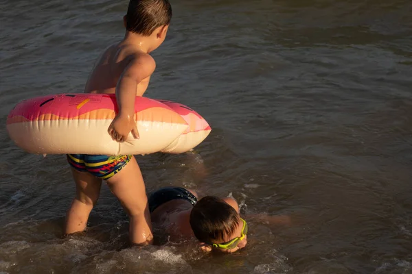 5 yaşındaki bir çocuk 10 yaşındaki abisiyle donut şeklinde şişirilebilir bir çemberle denizde yüzüyor. İki kardeş suya dalar.