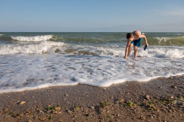 10 yaşındaki bir çocuk deniz kıyısında suyun içinde duran plaj terliklerini giyer..
