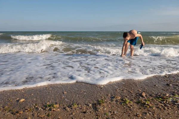 10 yaşındaki bir çocuk deniz kıyısında suyun içinde duran plaj terliklerini giyer..