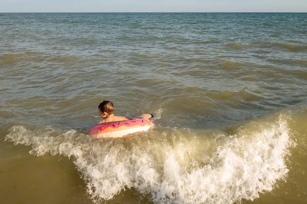Ένα χαρούμενο αγόρι 5 ετών κολυμπάει στη θάλασσα σε ένα φουσκωτό δαχτυλίδι με τη μορφή ενός ντόνατ σε μια ηλιόλουστη μέρα. — Φωτογραφία Αρχείου