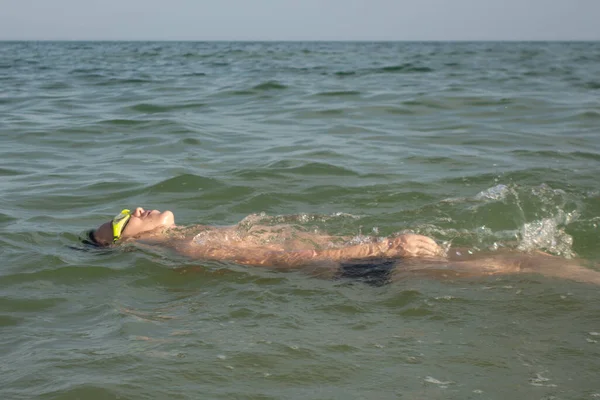 Boy 10 years old swims on his back in green swimming goggles in the sea