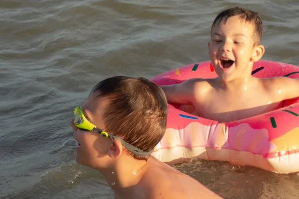 5 ve 10 yaşındaki kardeşler kıyıya yakın denizde yüzüp eğleniyorlar. Donut şeklinde şişme dairesi olan küçük çocuk, yeşil yüzme gözlüklü olan.