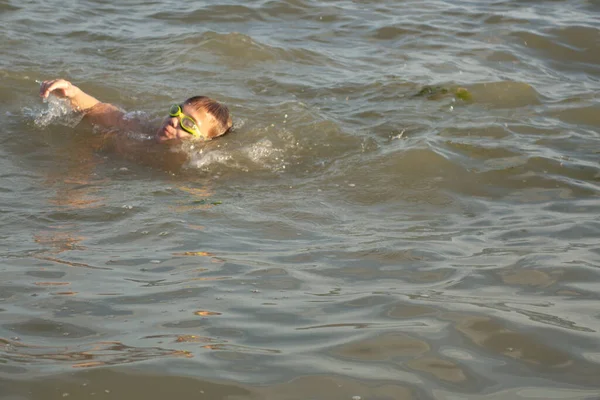 10 yaşında bir çocuk yeşil yüzme gözlükleriyle deniz kenarında yüzüyor ve eğleniyor.