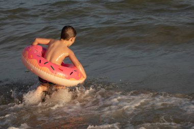 5 yaşında bir çocuk deniz kenarında yüzüyor ve şişme donut şeklinde bir çemberle eğleniyor.