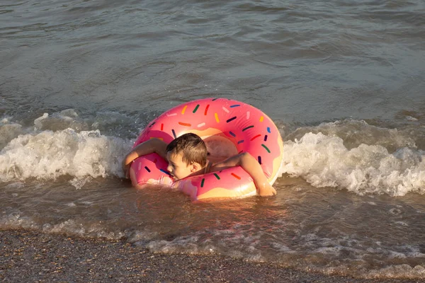 5 yaşında bir çocuk deniz kenarında yüzüyor ve şişme donut şeklinde bir çemberle eğleniyor.