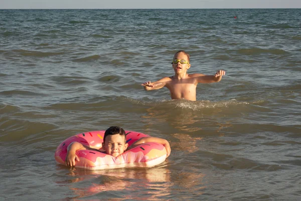 Brothers 5 and 10 years old swim and have fun in the sea near the shore. The younger boy with a donut-shaped inflatable circle, the older one in green swimming goggles