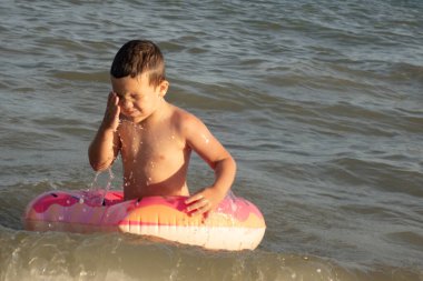 5 yaşında bir çocuk deniz kenarında yüzüyor ve şişme donut şeklinde bir çemberle eğleniyor.