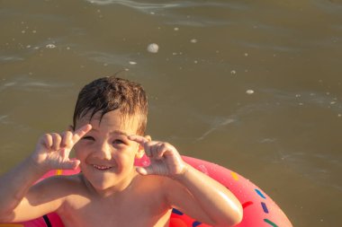5 yaşında bir çocuk denizde yüzüyor, seviniyor ve çörek şeklinde şişirilebilir bir yüzükle eğleniyor.