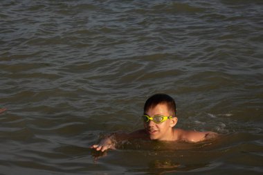 Yeşil yüzme gözlükleri takmış 10 yaşında bir çocuk kıyıya yakın denizde yüzüyor.