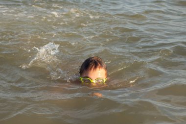 Yeşil yüzme gözlükleri takmış 10 yaşında bir çocuk kıyıya yakın denizde yüzüyor.