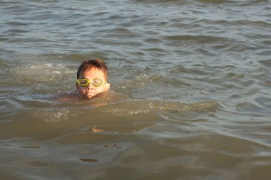 Yeşil yüzme gözlükleri takmış 10 yaşında bir çocuk kıyıya yakın denizde yüzüyor.