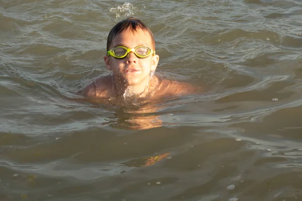 Yeşil yüzme gözlükleri takmış 10 yaşında bir çocuk kıyıya yakın denizde yüzüyor.