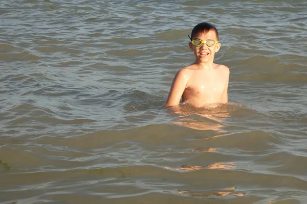 A 10-year-old boy in green swimming goggles swims in the sea near the shore