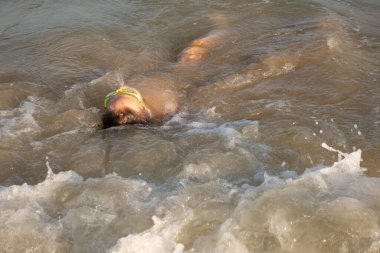 Yeşil yüzme gözlükleri takmış 10 yaşında bir çocuk kıyıya yakın denizde yüzüyor.