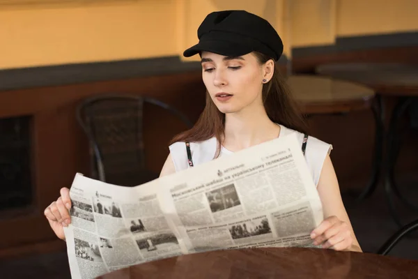 Young woman having a breakfast reading newspaper outdoors at the typical french cafe terrace in France. Playful girl with perfect long hairs posing outdoor. Wearing wool cap. Street fashion look. Cute teen girl.