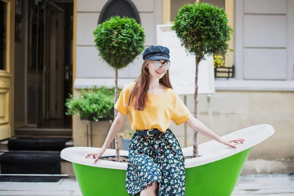 Oynak kız açık poz mükemmel uzun tüyleri ile. Sarı tişört giyen, Etek, güneş gözlüğü, yün kap. Sokak moda bakmak. Yeşil bath yakınındaki sevimli genç kız.