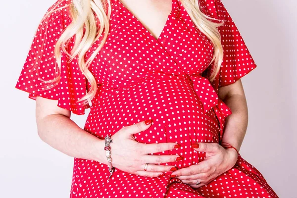 Pregnant woman in dress holds hands on belly on a white background. Pregnancy, maternity, preparation and expectation concept. Close-up, copy space, indoors. Beautiful tender mood photo of pregnancy.