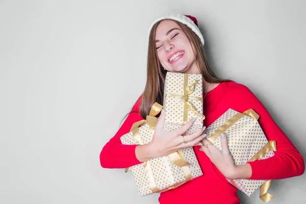Smiling teenage girl in santa helper hat with many gift boxes on white background. Positive emotional Santa girl. Concept of selling and Christmas shopping. Xmas. Girl hugging gifts.