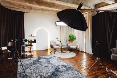 İyi aydınlatılmış bir stüdyoda, bir podcast kaydı için davetiye düzenlenir. İki rahat sandalye, mikrofon ve kameralarla donatılmış yuvarlak bir masaya bakıyor. Görüşmeler için sıcak bir ortam yaratıyor..