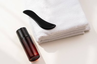 Siyah gua sha aleti, parlak bir banyo ortamında düzgünce katlanmış beyaz bir havlunun üzerinde, içi cilt ürünleriyle dolu koyu cam bir kapta duruyor..