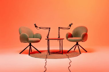 Two cozy chairs sit facing each other with microphones mounted on adjustable arms. The warm orange backdrop creates an inviting atmosphere for an engaging podcast recording session.
