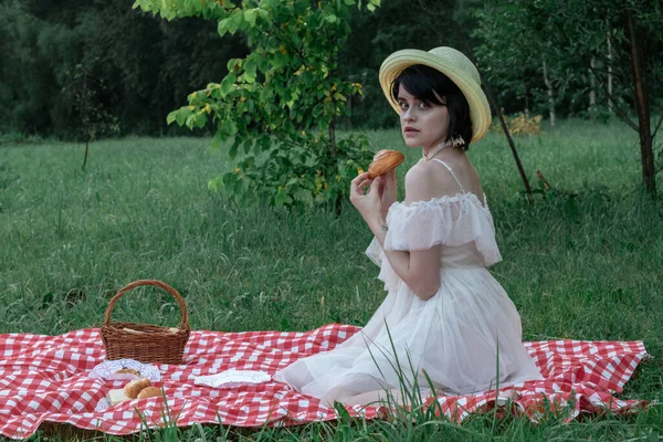 Красивая романтическая молодая женщина с темными короткими волосами в соломенной шляпе и белом платье сидит на пикнике, в природе — стоковое фото