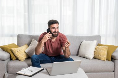 Genç sakallı, serbest çalışan bir adam kanepede oturuyor telefonda konuşuyor ve dizüstü bilgisayarı üzerinde çalışıyor yüzünde gülümsemeyle.