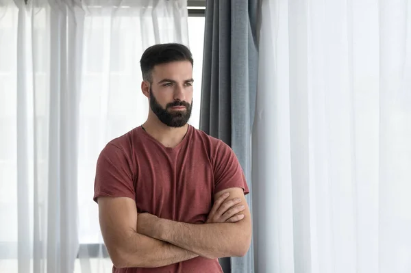 Young handsome bearded depressed man standing alone and looking through the window felling depression and anxiety after his girlfriend left him