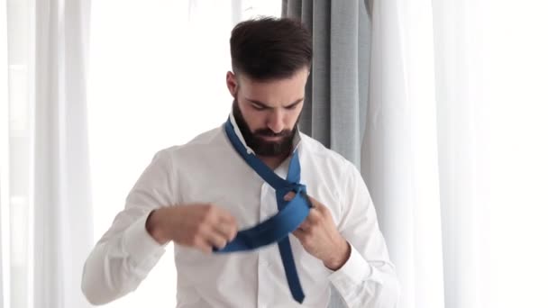 Junge bärtige gutaussehende Mann in weißem Hemd versucht, seine Krawatte Krawatte Krawatte Vorbereitung für das erste Vorstellungsgespräch oder Date und haben Problem, weil es sein erstes Mal ist und es kompliziert ist