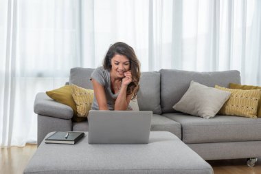 Genç ve güzel bekar bir kadın dizüstü bilgisayarında erkek arkadaşı veya kız arkadaşıyla sohbet ediyor koltukta oturuyor ve büyük bir gülümsemeyle gülüyor.