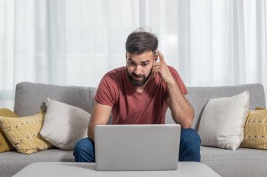 Genç, sakallı, serbest çalışan, kanepenin üzerinde oturmuş, bir müşterisiyle dizüstü bilgisayardan şirketinin yeni iş projesini konuşuyor.