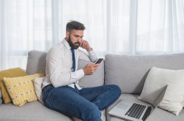 Genç serbest çalışan işadamı evinde kanepede oturuyor ve patronuna serbest işçi olarak yaptığı yeni iş projesini göstermek için telefon ve dizüstü bilgisayarla video konferansı bekliyor. 