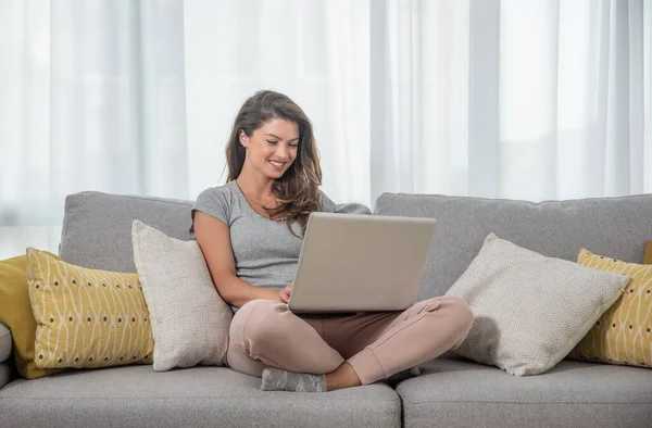 Genç ve güzel bekar bir kadın dizüstü bilgisayarında erkek arkadaşı veya kız arkadaşıyla sohbet ediyor koltukta oturuyor ve büyük bir gülümsemeyle gülüyor.