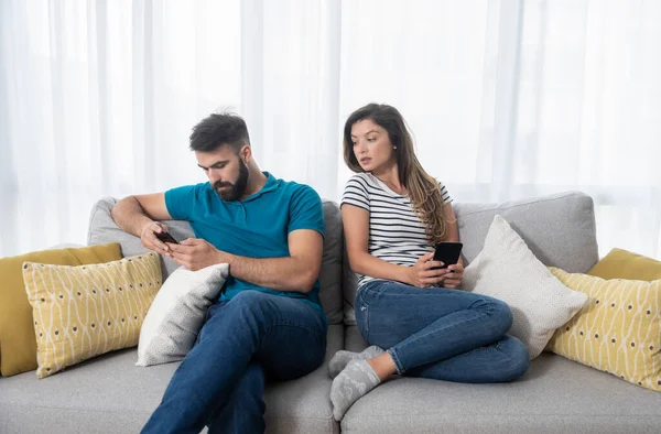 Stock image Young boyfriend making fun and jealous his girlfriend pretending that he is texting with other girl on his cell or mobile phone while they sitting on the sofa in their apartment