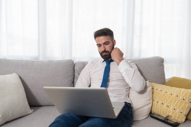 Genç, sakallı, serbest çalışan, kanepenin üzerinde oturmuş, bir müşterisiyle dizüstü bilgisayardan şirketinin yeni iş projesini konuşuyor.