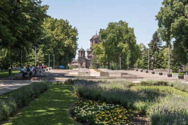 Sırbistan 'ın başkenti Belgrad' daki Tasmajdan parkının manzarasında banklarda oturan insanlar ve uzaktaki Sırp Ortodoks Kilisesi Saint Marko. 