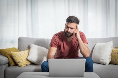 Genç, sakallı, serbest çalışan, kanepenin üzerinde oturmuş, bilgisayarla müşterisi için ev konseptinden yeni bir iş projesiyle ilgili konuşuyor.