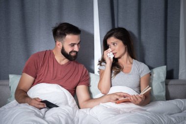 Genç adam, kız arkadaşı yatakta kitap okuyup ağlarken ağladığı için gülüp eğleniyor.