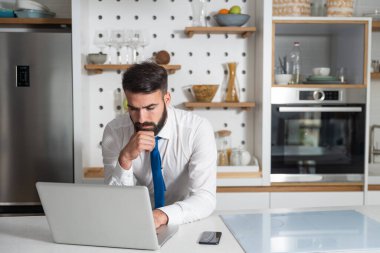Genç sakallı işadamı mutfakta duruyor ve yeni iş planı için şirket iş listesini kontrol ediyor ve dizüstü bilgisayarında projesini yapıyor.