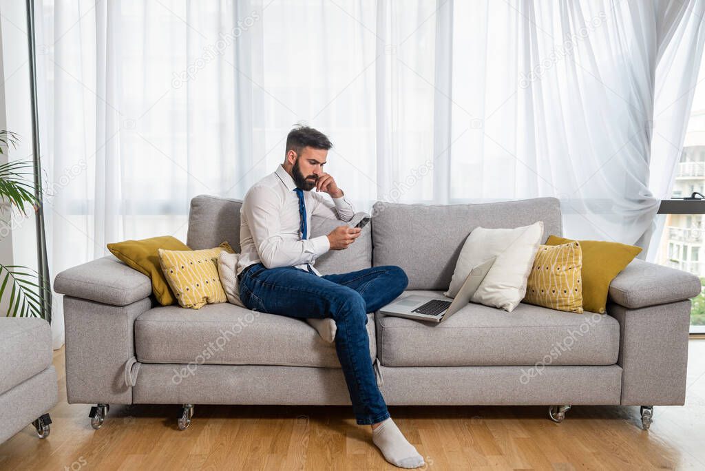 Young freelancer business man sitting on sofa at his home and taking a break on his phone of laptop and waiting for his boss to talk about new business project as freelance worker 