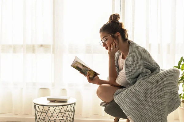 Young beautiful girl with reading glasses sitting in the chair near window reading the book with natural sunlight and enjoy, with film grain
