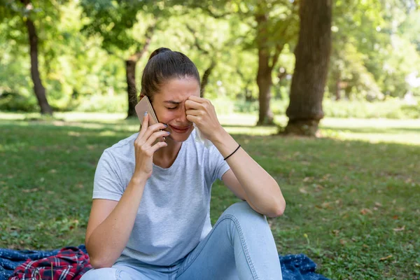年轻悲伤的女人坐在公园里拿着手机哭泣 因为她得到了一个坏的新东西 — 图库照片