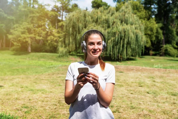 Girl with wireless headphones and phone standing in the park listening the music