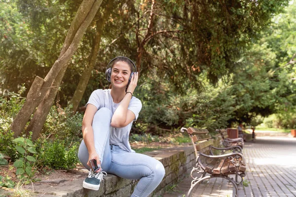 Genç ve güzel bir kadın kafasında kablosuz kulaklıkla parkta oturuyor ve müzik dinliyor.