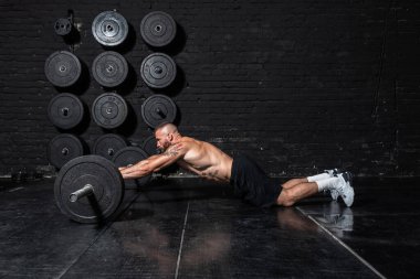 Genç ve güçlü, kaslı, terli, kaslı bir adam spor salonunun zemininde halter sallayarak vücut çalışıyor.