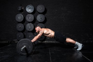 Genç ve güçlü, kaslı, terli, kaslı bir adam spor salonunun zemininde halter sallayarak vücut çalışıyor.