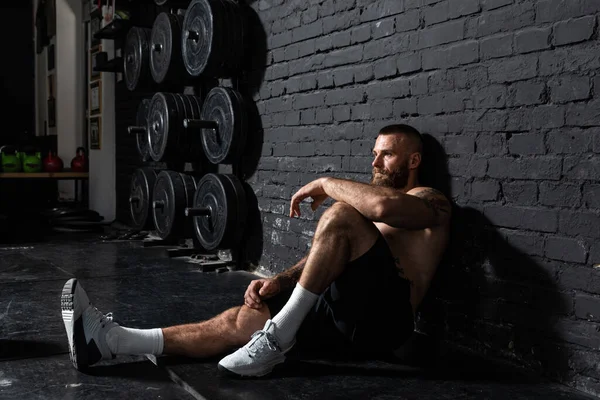 Genç, güçlü, terli, formda, kaslı adam spor salonunda oturuyor ve vücut çalışmalarına ara veriyor.