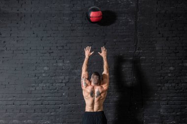 Genç, güçlü, terli, formda, kaslı, iri kaslı bir adam spor salonunda sıkı antrenman yapmak için duvarlara ilaç topu atıyor.