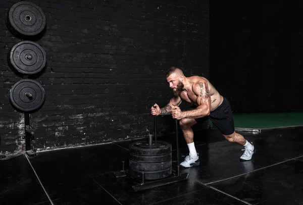 stock image Young strong sweaty muscular fit man with big muscles pushing heavy weight plates on the prowler slider in the gym as hardcore cross workout training