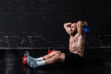 Genç, terli, güçlü, kaslı, iri kaslı bir adam spor salonunun zemininde oturuyor ve ağır kettlebell ile triseps antrenmanı yapıyor.
